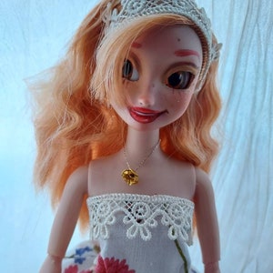 Op de afbeelding: Een pop met lang, krullend, oranje haar die een witte jurk draagt met een kanten rand en een gouden bloemenketting. De pop glimlacht en heeft een hoofdband met kant op.