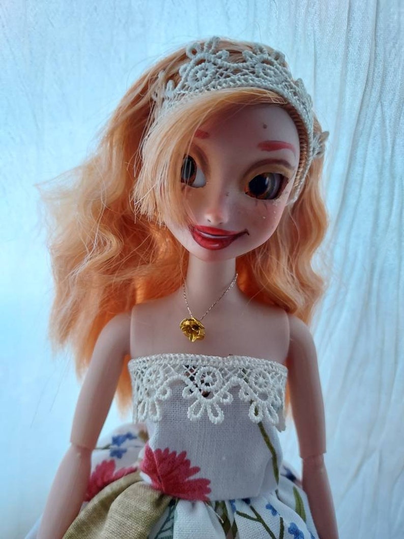 Op de afbeelding: Een pop met lang, krullend, oranje haar die een witte jurk draagt met een kanten rand en een gouden bloemenketting. De pop glimlacht en heeft een hoofdband met kant op.