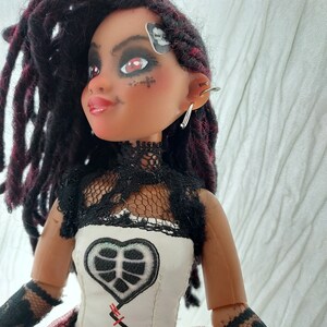 May include: A doll with dark red and black dreadlocks, wearing a white and black lace top with a black heart-shaped patch and red stitching. The doll has red eyeshadow and a black skull sticker on her forehead.