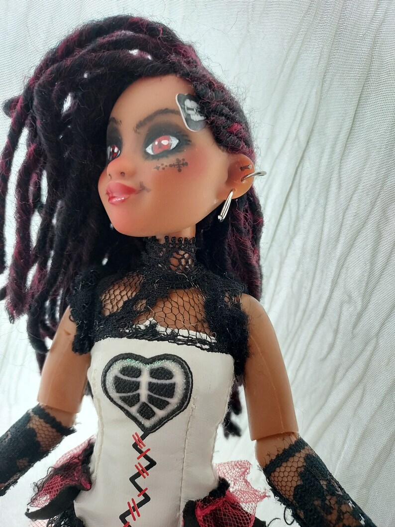 May include: A doll with dark red and black dreadlocks, wearing a white and black lace top with a black heart-shaped patch and red stitching. The doll has red eyeshadow and a black skull sticker on her forehead.