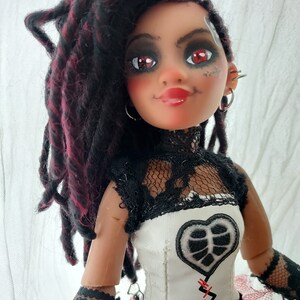 May include: A doll with red and black dreadlocks, wearing a white and black lace dress with a heart-shaped patch and red stitching. The doll has red eyeshadow and red lipstick.