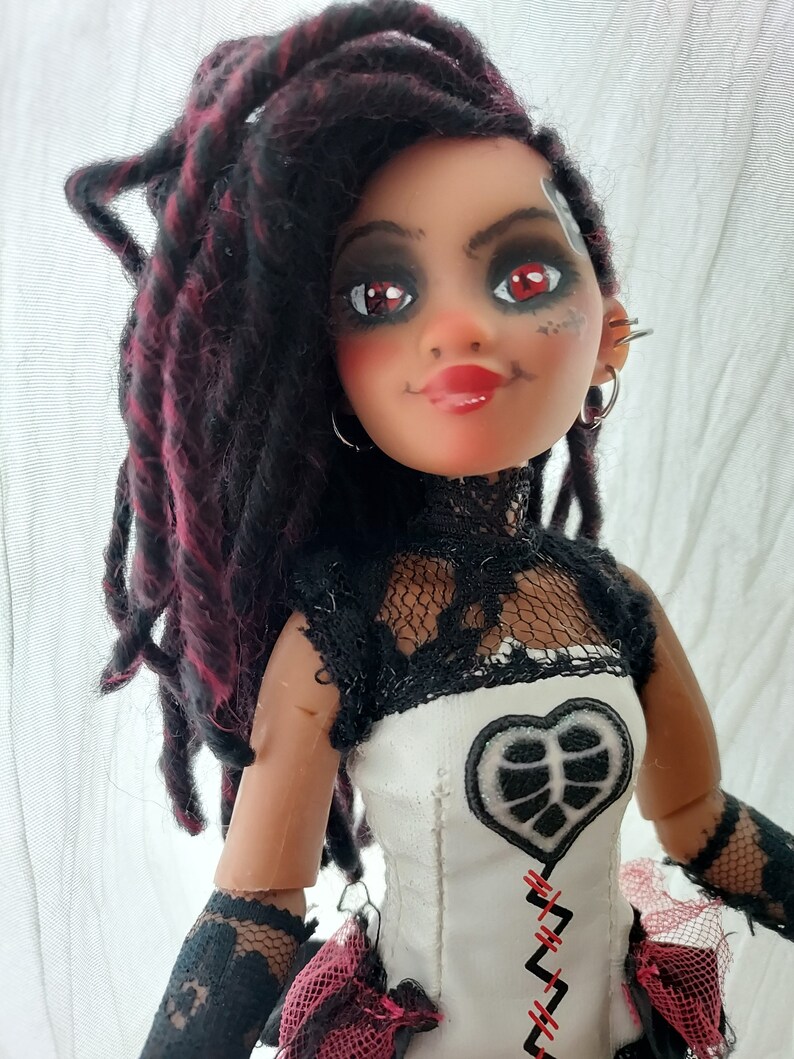 May include: A doll with red and black dreadlocks, wearing a white and black lace dress with a heart-shaped patch and red stitching. The doll has red eyeshadow and red lipstick.