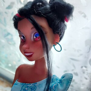 May include: A doll with black hair styled in two buns, wearing a blue and white dress with a cartoon bear print. The doll has blue eyes and is wearing turquoise hoop earrings.