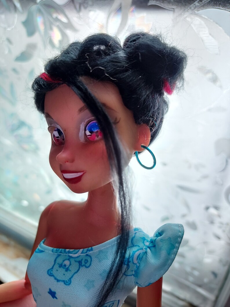 May include: A doll with black hair styled in two buns, wearing a blue and white dress with a cartoon bear print. The doll has blue eyes and is wearing turquoise hoop earrings.