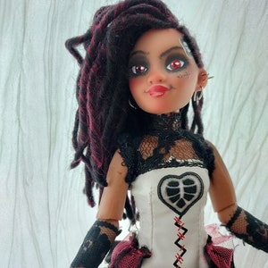May include: A doll with dark brown dreadlocks with red streaks, wearing a white dress with black lace and a red heart-shaped patch. The doll has red eyeshadow and red lipstick.