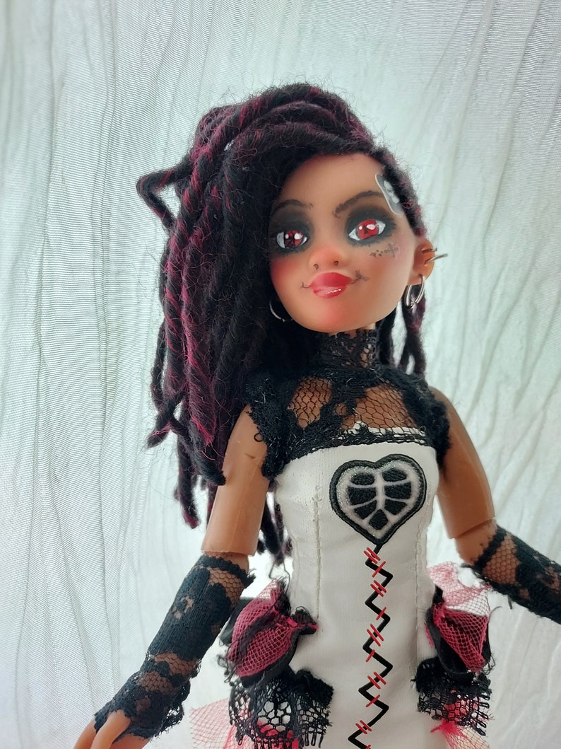 May include: A doll with dark brown dreadlocks with red streaks, wearing a white dress with black lace and a red heart-shaped patch. The doll has red eyeshadow and red lipstick.