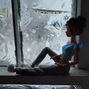 May include: A doll with dark brown hair and a blue and white top sits on a windowsill with a floral patterned window film behind her. She is wearing gray pants and white fuzzy slippers.