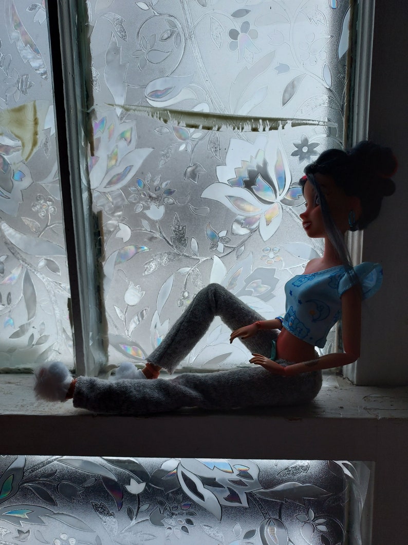May include: A doll with dark brown hair and a blue and white top sits on a windowsill with a floral patterned window film behind her. She is wearing gray pants and white fuzzy slippers.
