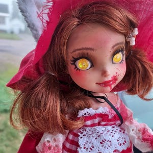 Könnte beinhalten: Eine Puppe mit roter Kapuze, weißem Pelzbesatz, rot-weißem kariertem Kleid und weißem Spitzenbesatz. Die Puppe hat rote Haare, gelbe Augen und einen blutigen Mund.