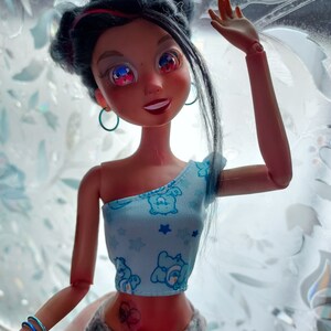 May include: A doll with long black hair, wearing a blue and white top with a cartoon bear print and grey pants. The doll has a heart-shaped tattoo on her stomach and is wearing blue earrings.