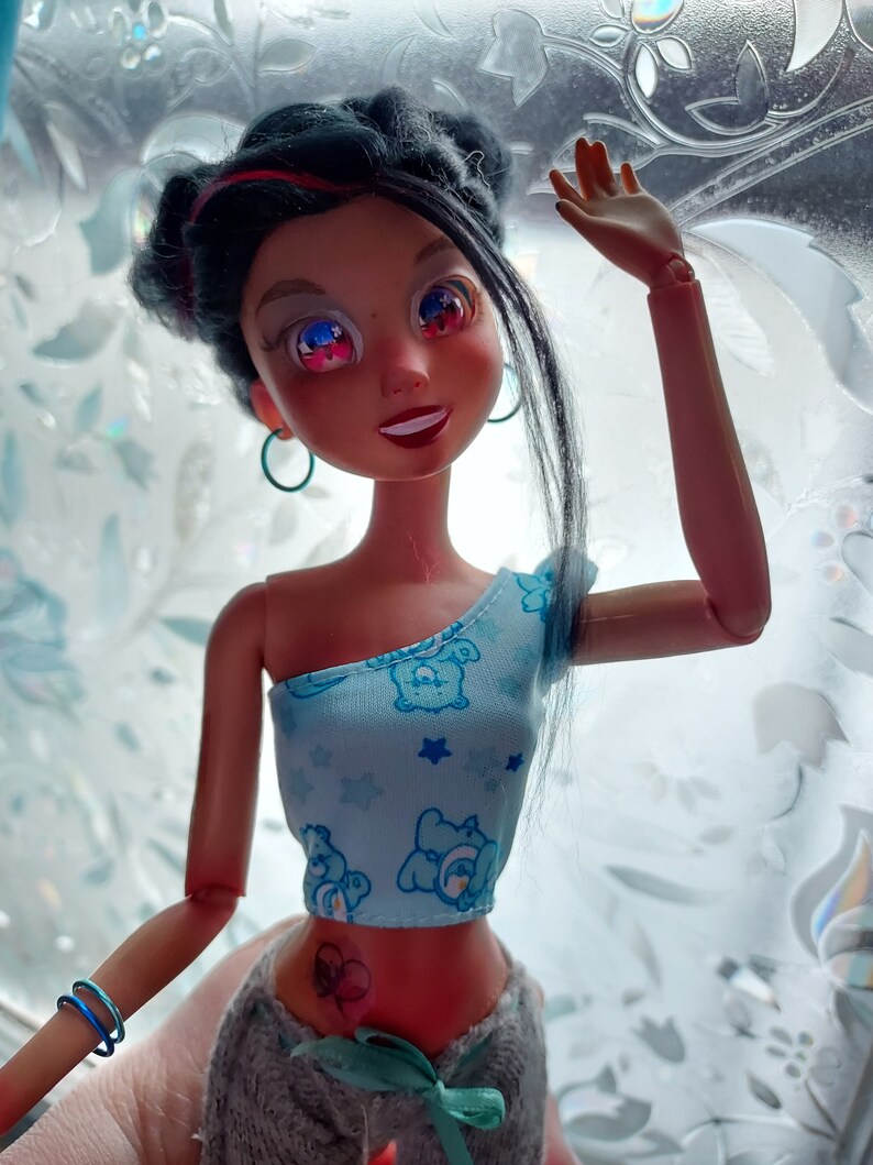 May include: A doll with long black hair, wearing a blue and white top with a cartoon bear print and grey pants. The doll has a heart-shaped tattoo on her stomach and is wearing blue earrings.