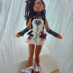 May include: A doll with long black dreadlocks, wearing a white dress with black lace trim and red accents, and red and white sneakers.
