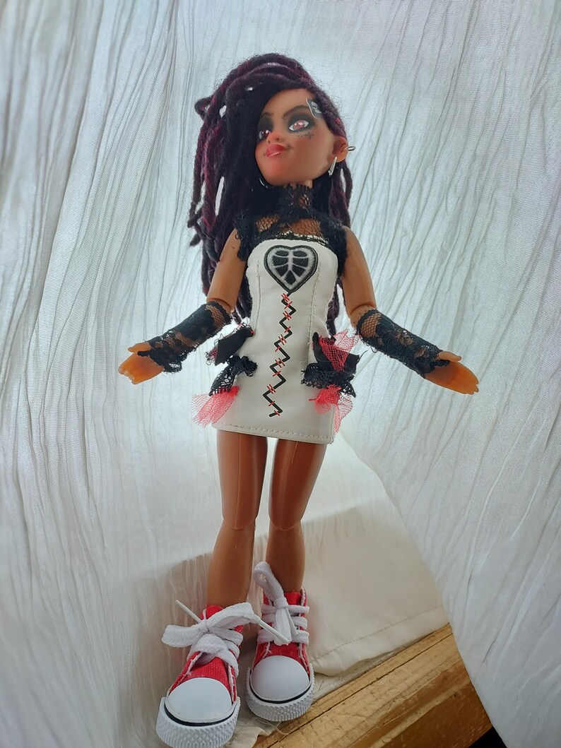 May include: A doll with long black dreadlocks, wearing a white dress with black lace trim and red accents, and red and white sneakers.