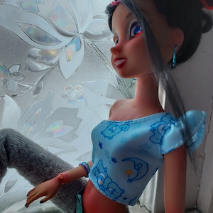 May include: A doll with long black hair wearing a blue crop top with a cartoon bear print and gray pants. The doll has blue eyes and red lipstick.