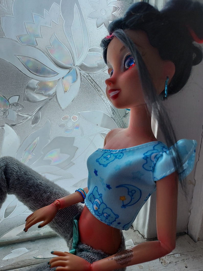 May include: A doll with long black hair wearing a blue crop top with a cartoon bear print and gray pants. The doll has blue eyes and red lipstick.