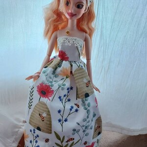 Op de afbeelding: Een pop die een witte jurk draagt met een bloemen- en bijenkorfpatroon. De jurk heeft een kanten rand en de pop draagt een witte kanten kroon.