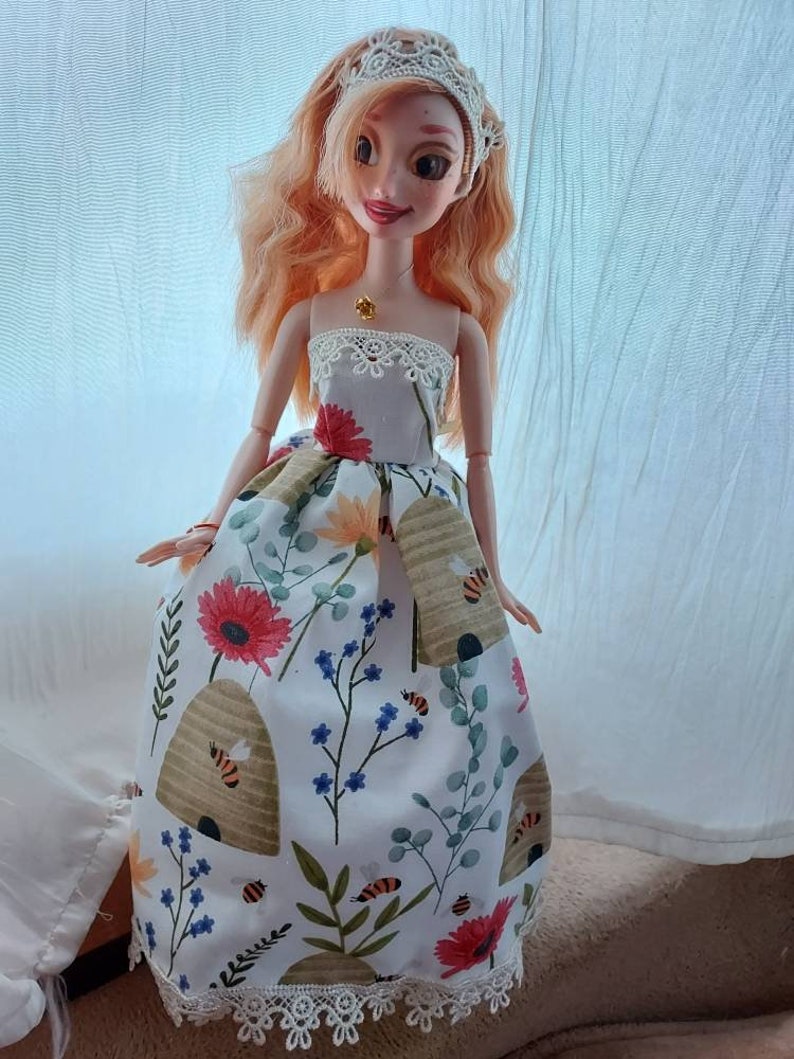 Op de afbeelding: Een pop die een witte jurk draagt met een bloemen- en bijenkorfpatroon. De jurk heeft een kanten rand en de pop draagt een witte kanten kroon.