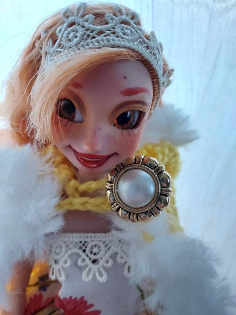 Op de afbeelding: Een pop met blond haar die een witte kanten kroon, een gele gebreide sjaal en een witte bontjas draagt. De pop heeft een parel en gouden broche.