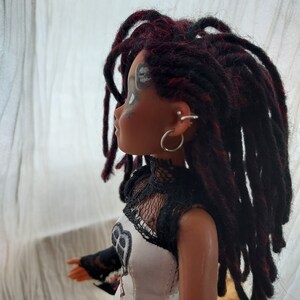 May include: A doll with long black and red dreadlocks, wearing a white and black corset-style top with lace detailing. The doll has black eyeliner and a silver hoop earring.