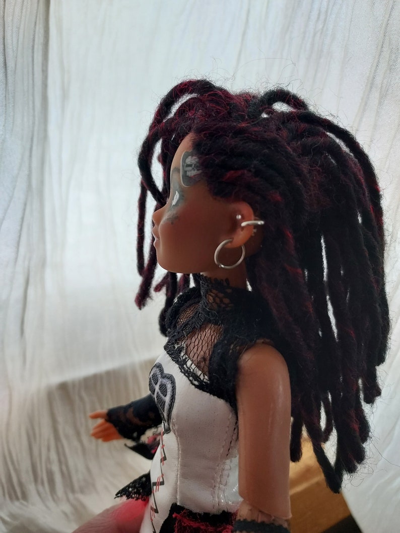 May include: A doll with long black and red dreadlocks, wearing a white and black corset-style top with lace detailing. The doll has black eyeliner and a silver hoop earring.