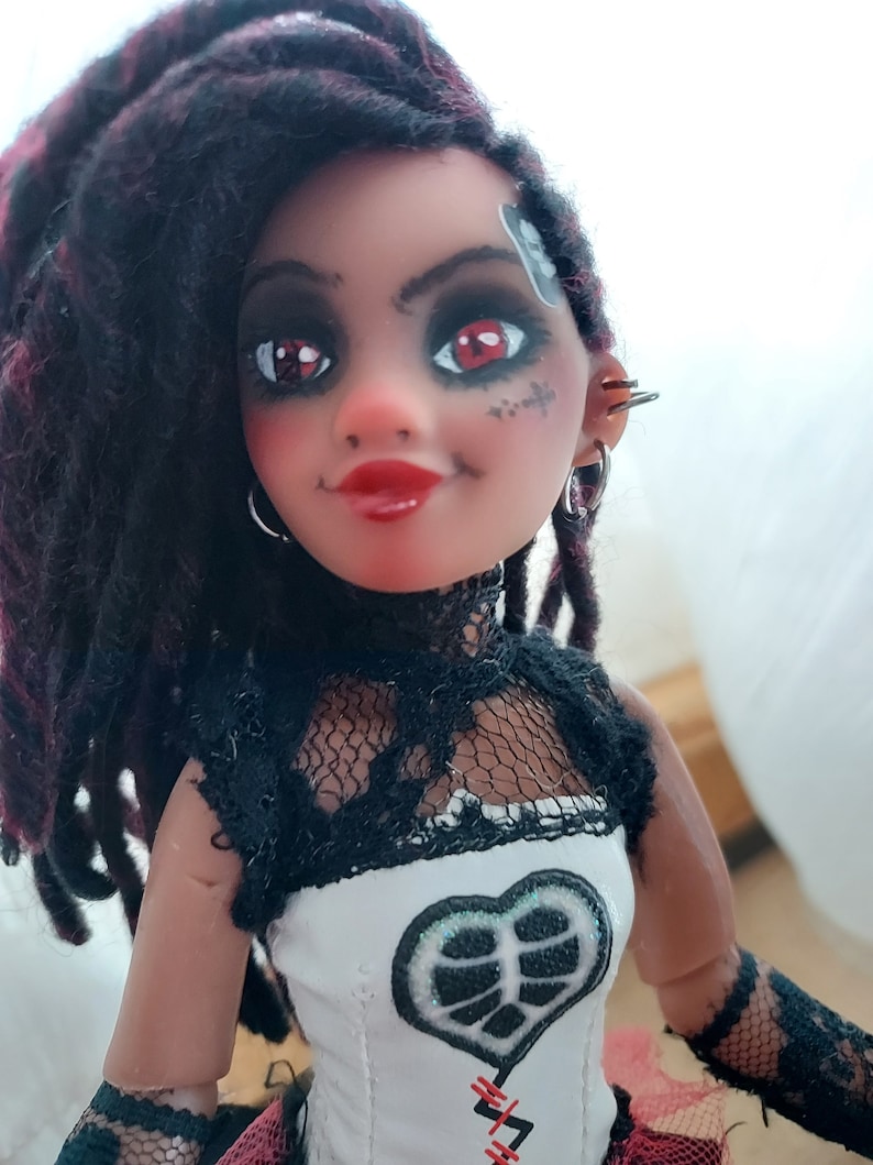 May include: A doll with dark red dreadlocks, black and white lace clothing, and a white corset with a black heart design. The doll has red eyeshadow and a black and white tattoo on her face.