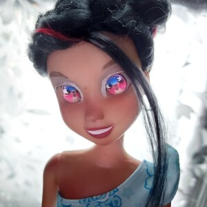 May include: A doll with black hair and a blue and white patterned dress. The doll has pink and blue eyes with a white background.