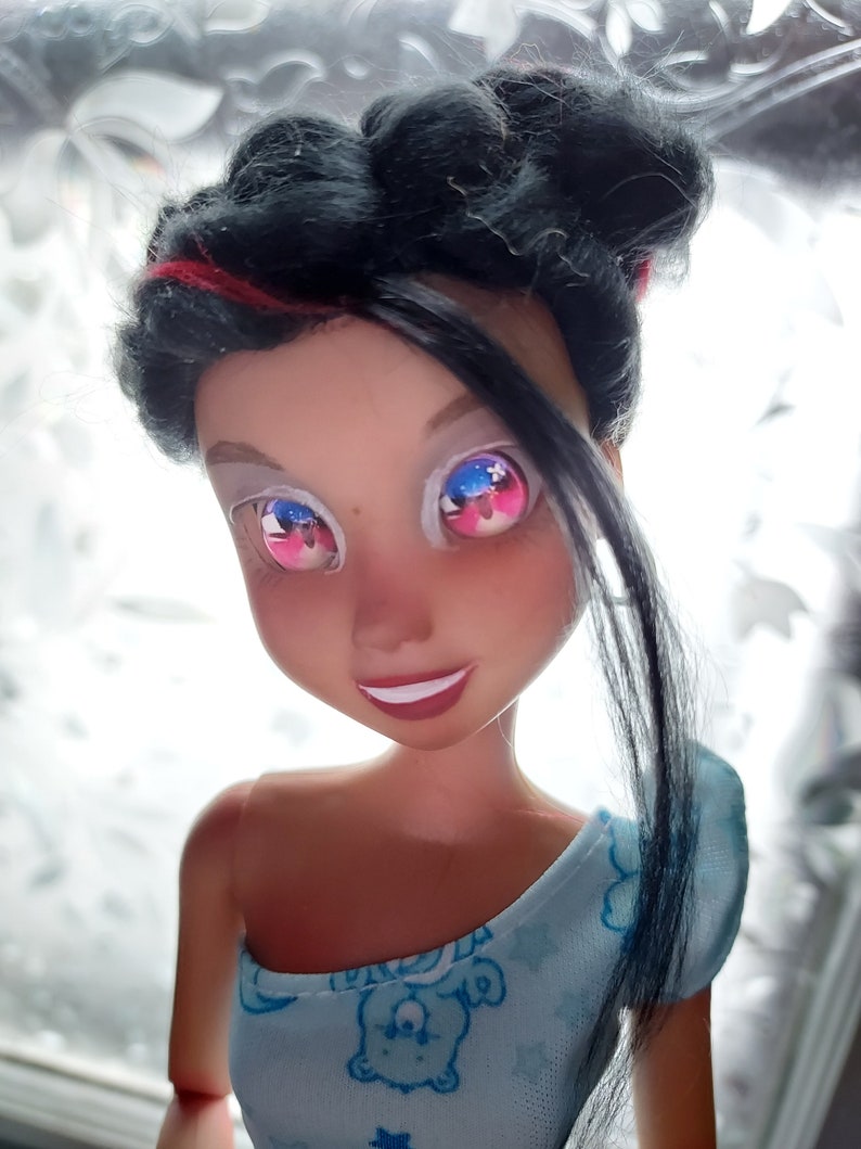 May include: A doll with black hair and a blue and white patterned dress. The doll has pink and blue eyes with a white background.