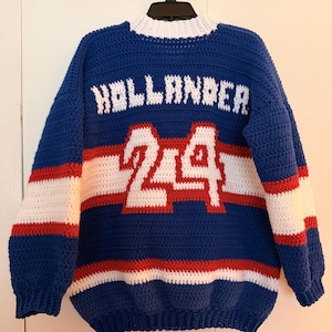 May include: A blue, red, and white crocheted sweater with the name "HOLLANDER" and the number "24" on the front. The sweater has a white collar and cuffs, with red and white stripes across the chest and sleeves. The sweater is hanging on a black hanger.
