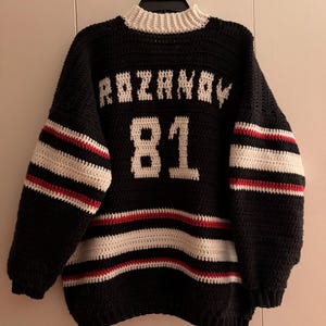Può includere: Una maglia da hockey all'uncinetto in bianco e nero con il nome "ROZANOV" e il numero "81" in bianco. La maglia presenta strisce bianche e rosse sulle maniche e un colletto bianco.