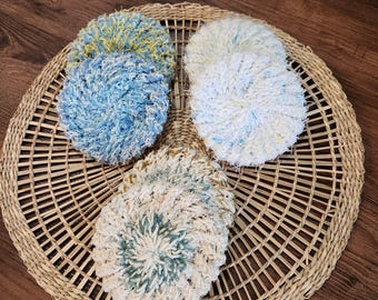 May include: A collection of handmade, round, textured scrubbies in various colors, including blue, yellow, and white. The scrubbies are arranged on a woven, circular placemat, with a wooden background.