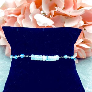 Puede incluir: Un brazalete delicado con cuentas de color azul claro y blanco en una cadena de plata. El brazalete se muestra en un cojín de terciopelo negro.