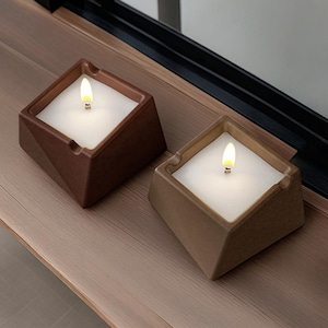 Puede incluir: Dos velas cuadradas marrones con un diseño geométrico. Cada vela tiene un interior de cera blanca y una pequeña llama parpadeante. Las velas están colocadas sobre una superficie de madera.