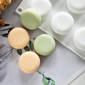 May include: A silicone mold for making round soap bars. The mold has six cavities and is shown with three finished soap bars in green, light orange, and white.