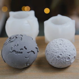 Op de afbeelding: Twee grijze betonnen bollen, een met een gedetailleerde kaart van de aarde en de andere met een met kraters bedekt oppervlak dat lijkt op de maan. Twee witte siliconen mallen zijn ook te zien, waarschijnlijk gebruikt om de bollen te maken.