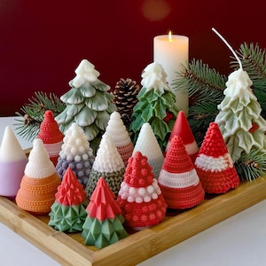 May include: A selection of Christmas tree-shaped candles in shades of green, red, white, and orange. Some candles have textured surfaces, others are smooth. A lit white pillar candle and pine needles are visible in the background, adding to the festive theme.