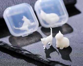 Op de afbeelding: Twee witte keramische kattenbeeldjes, een zittend en een staand, op een zwart oppervlak. De beeldjes bevinden zich in doorzichtige plastic containers.