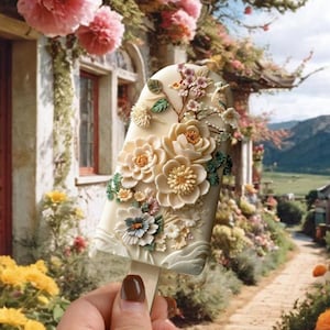 Może przedstawiać: Biała czekoladowa lizak ozdobiona misternymi kwiatami z cukru w odcieniach bieli, różu i zieleni. Lizak jest trzymany w dłoni na tle ogrodu z białym budynkiem i ścieżką.