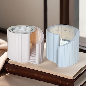 May include: Two decorative candle holders. One is white with a silver rim and white stones, the other is light blue with a lit candle inside. Both have a curved, ribbed design and sit on a book.