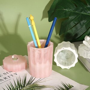 May include: A pink, textured, cylindrical container with a lid. The container has three pencils inside, one yellow, one blue, and one light blue. The container is sitting on a white surface with a book and a white, textured, cylindrical container with a lid.