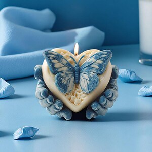 May include: A heart-shaped candle with a blue butterfly design and a lit wick. The candle is held by two hands and sits on a blue surface. Decorative blue petals are scattered around the candle.