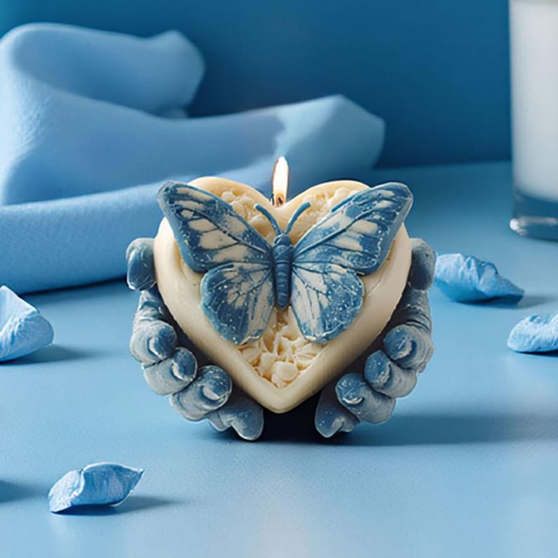 May include: A heart-shaped candle with a blue butterfly design and a lit wick. The candle is held by two hands and sits on a blue surface. Decorative blue petals are scattered around the candle.