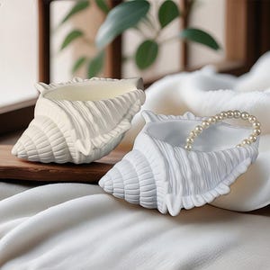 Könnte beinhalten: Zwei weiße, muschelförmige Dekorationsschalen. Eine ist leer, die andere enthält eine Perlenkette. Die Schalen haben eine strukturierte Oberfläche und sind auf einem weißen Stoff platziert. Die Schalen sind hellcremefarben.