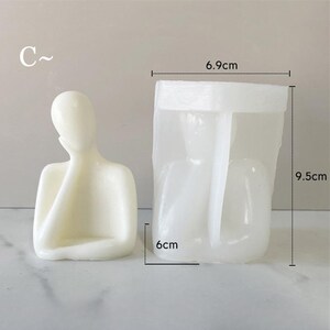 May include: A cream-colored candle in the shape of a person with their hand on their face, next to a clear plastic mold. The mold is rectangular, measuring 6 cm wide, 6.9 cm long, and 9.5 cm tall. The letter "C" is in the upper left corner.