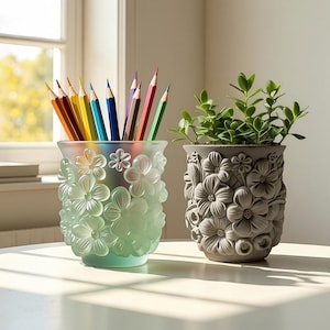 Puede incluir: Dos recipientes decorativos sobre una superficie blanca. Un portalápices de vidrio verde claro con flores en relieve contiene lápices de colores. Una maceta gris con un diseño floral contiene una planta verde.