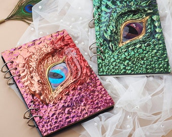 Può includere: Due quaderni a spirale con copertine iridescenti che presentano un designo a forma di occhio di drago. Un quaderno ha una copertina verde e l'altro una copertina rosa.