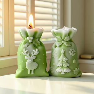 May include: Two green Christmas-themed candles shaped like gift bags. One candle has a reindeer design, the other a Christmas tree. Both have white accents and are lit with a warm flame. The candles are on a white surface.