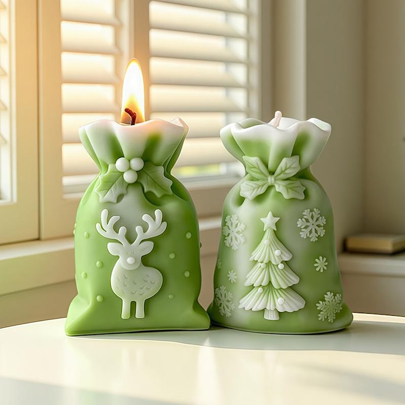 May include: Two green Christmas-themed candles shaped like gift bags. One candle has a reindeer design, the other a Christmas tree. Both have white accents and are lit with a warm flame. The candles are on a white surface.