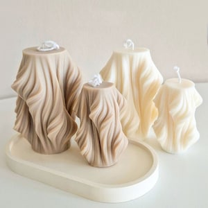 May include: Four decorative candles with a wavy, draped fabric design. The candles are in shades of beige and white and are arranged on a beige oval tray.