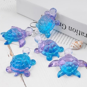 May include: Five translucent turtle figurines in shades of blue and purple are arranged on a white surface. The turtles have detailed shell patterns and flipper-like limbs. A seashell is visible in the background.