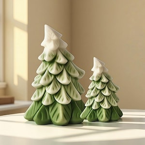 Puede incluir: Dos árboles de Navidad decorativos de cerámica. Los árboles son verdes con puntas blancas y tienen un diseño en capas en forma de hojas. El árbol más grande mide aproximadamente 20 cm de alto, y el más pequeño, unos 13 cm. Están colocados sobre una superficie blanca.